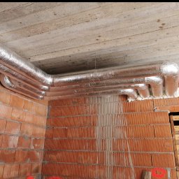 Aleksander Smoleń VentiPRO - Izolowane rury wentylacyjne pod betonowym stropem w surowym wnętrzu. Widoczne ceglane ściany i drabina. Detal z budowy, instalacja wentylacyjna w trakcie prac.