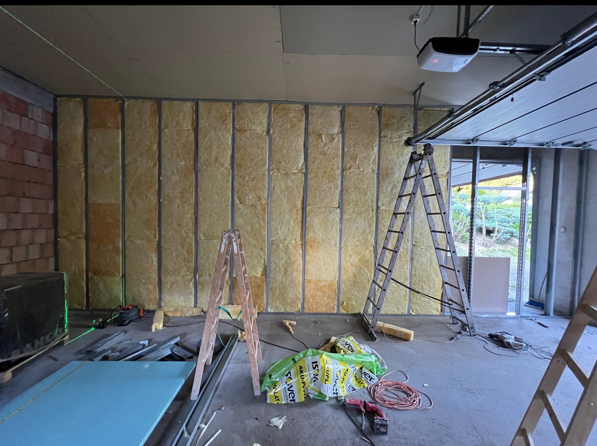 Ocieplanie ścian wełną mineralną w garażu z widocznymi elementami konstrukcyjnymi, narzędziami i workami z materiałem izolacyjnym. Widoczny laser krzyżowy.