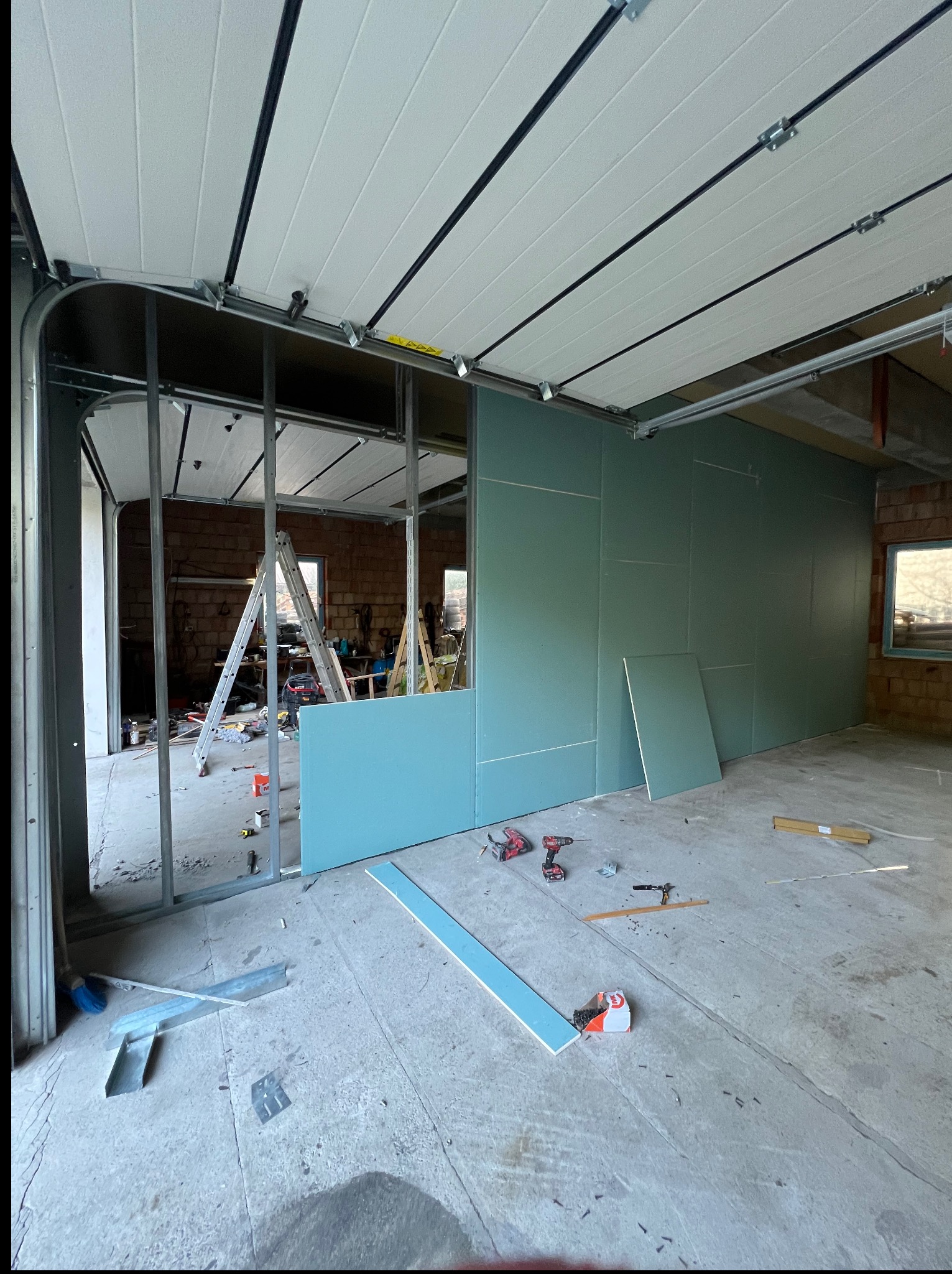 Montaż ścian działowych z płyt gipsowo-kartonowych w garażu. Widoczne profile, płyty w kolorze zielonym, narzędzia i drabina. Prace wykończeniowe w surowym wnętrzu.