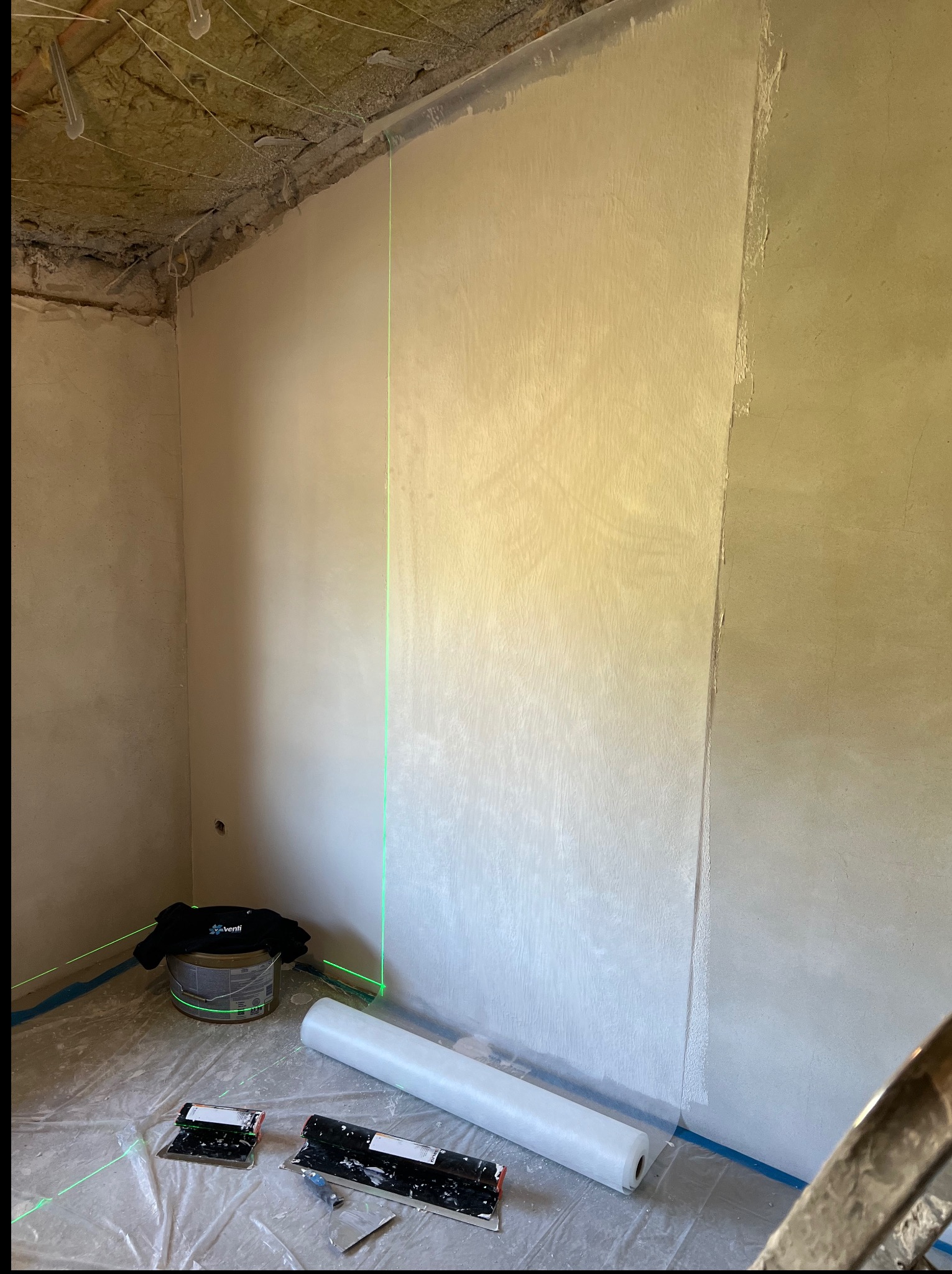 Ściana po nałożeniu gładzi, widoczny laser krzyżowy, narzędzia glazurnicze i wiadro z gładzią. Podłoga zabezpieczona folią malarską.