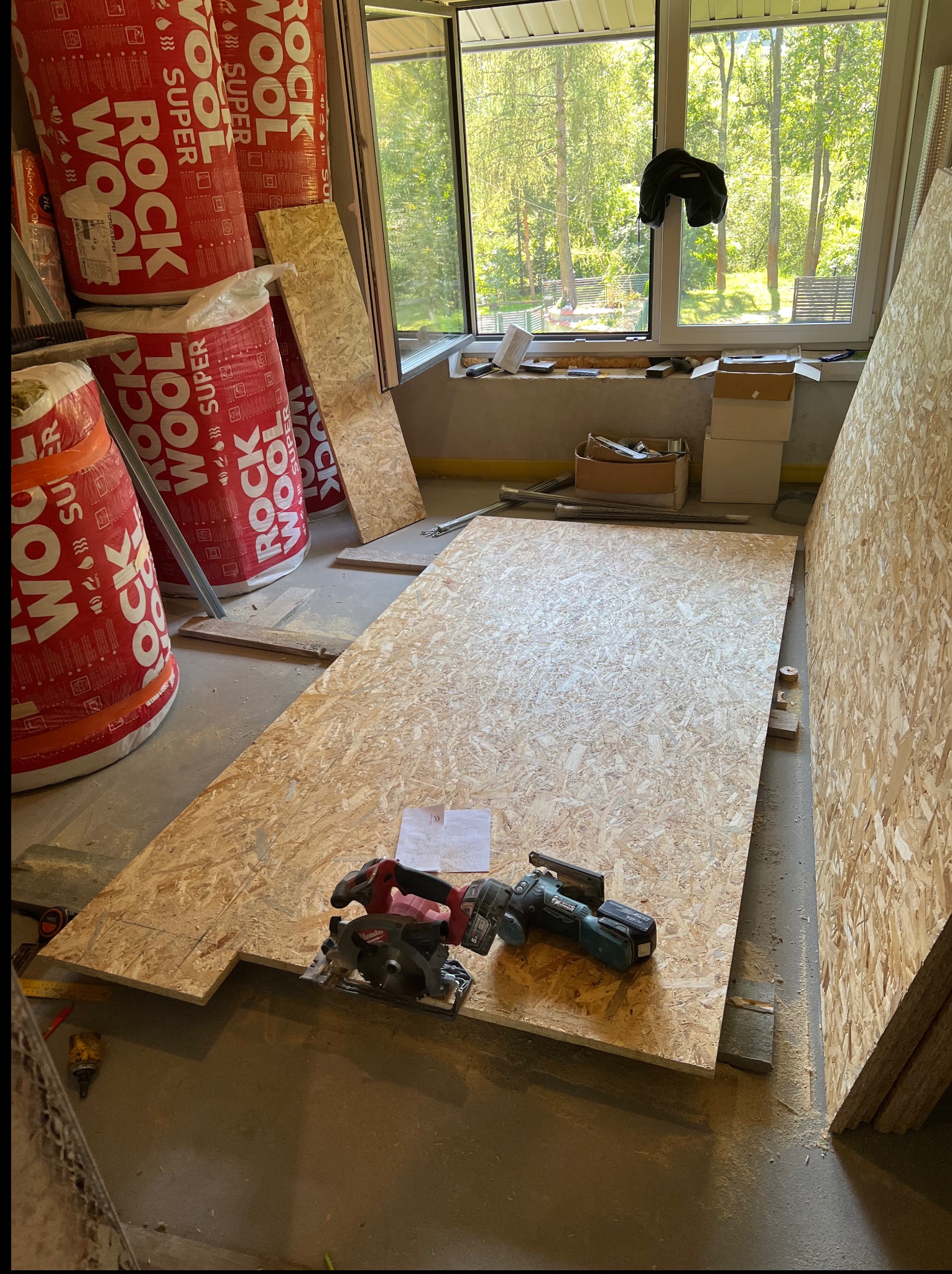 Przygotowanie do ocieplenia poddasza: płyta OSB, narzędzia i izolacja Rockwool Super Wool w pomieszczeniu z oknem na ogród. Etap prac wykończeniowych.