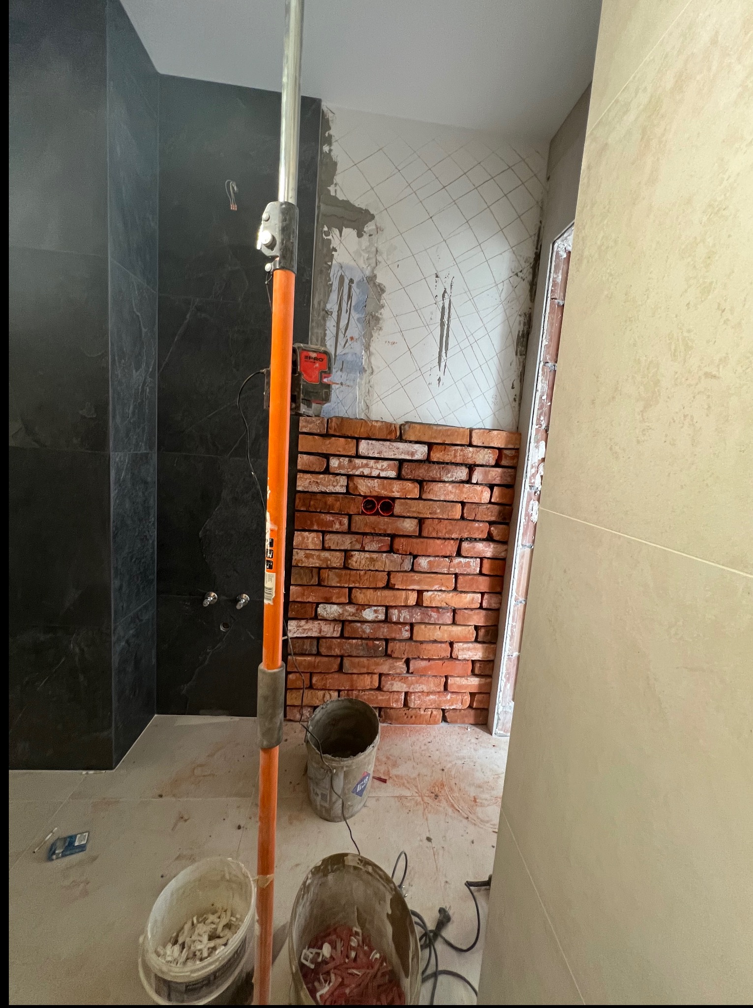 Remont łazienki: fragment ściany z cegieł, obok czarne płytki imitujące marmur. Widoczne narzędzia i wiaderka. Etap prac przed wykończeniem.