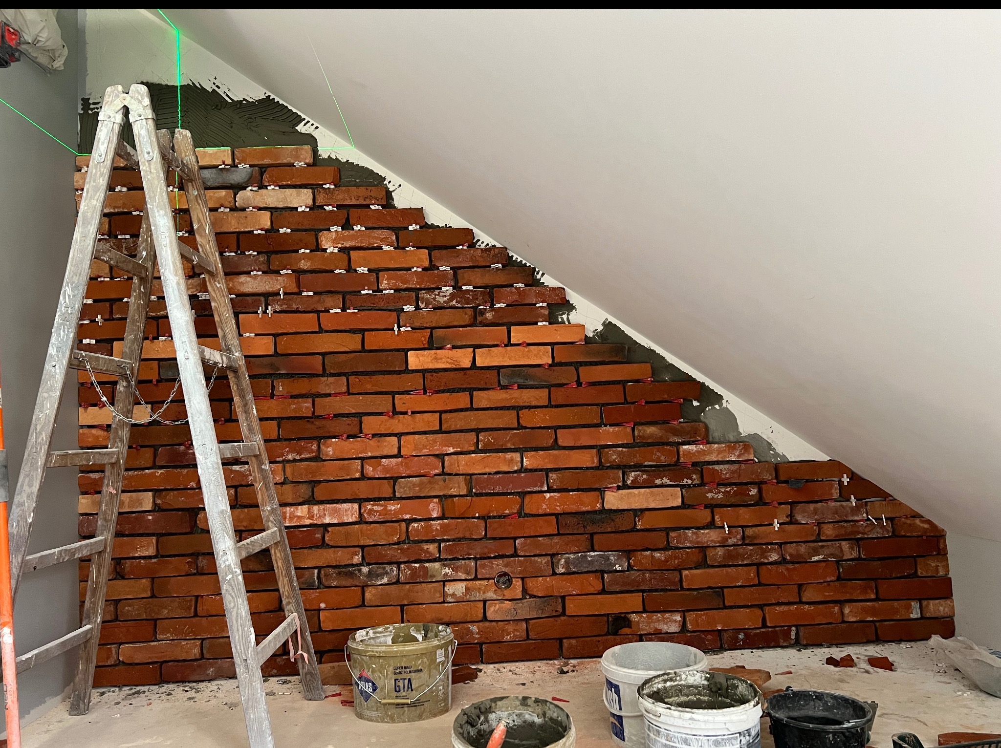 Układanie cegieł na ścianie pod skosem, widoczne poziomice laserowe, drabina i wiadra z zaprawą. Cegły w odcieniach czerwieni i brązu, białe fugi. Prace wykończeniowe.