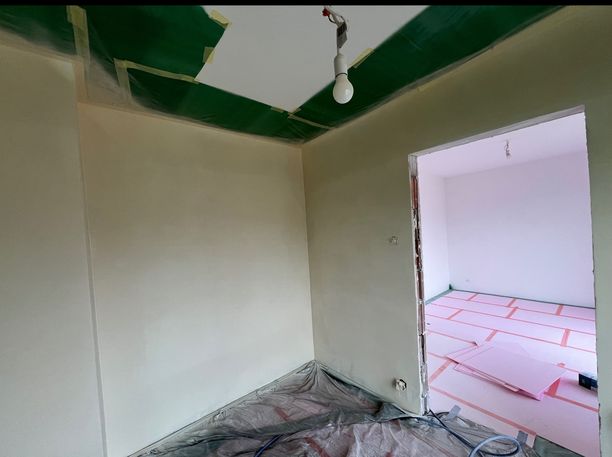 Świeżo pomalowane ściany w pokoju, zabezpieczone folią i taśmą malarską, z widocznym ociepleniem podłogi w sąsiednim pomieszczeniu. Żarówka zwisa z sufitu.