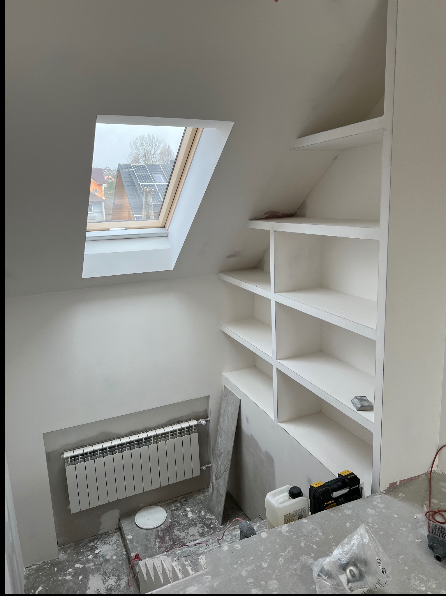 Białe, wnękowe półki w skosie poddasza, widok na okno dachowe i grzejnik. Ślady prac wykończeniowych na podłodze. Detal wnętrza po remoncie.