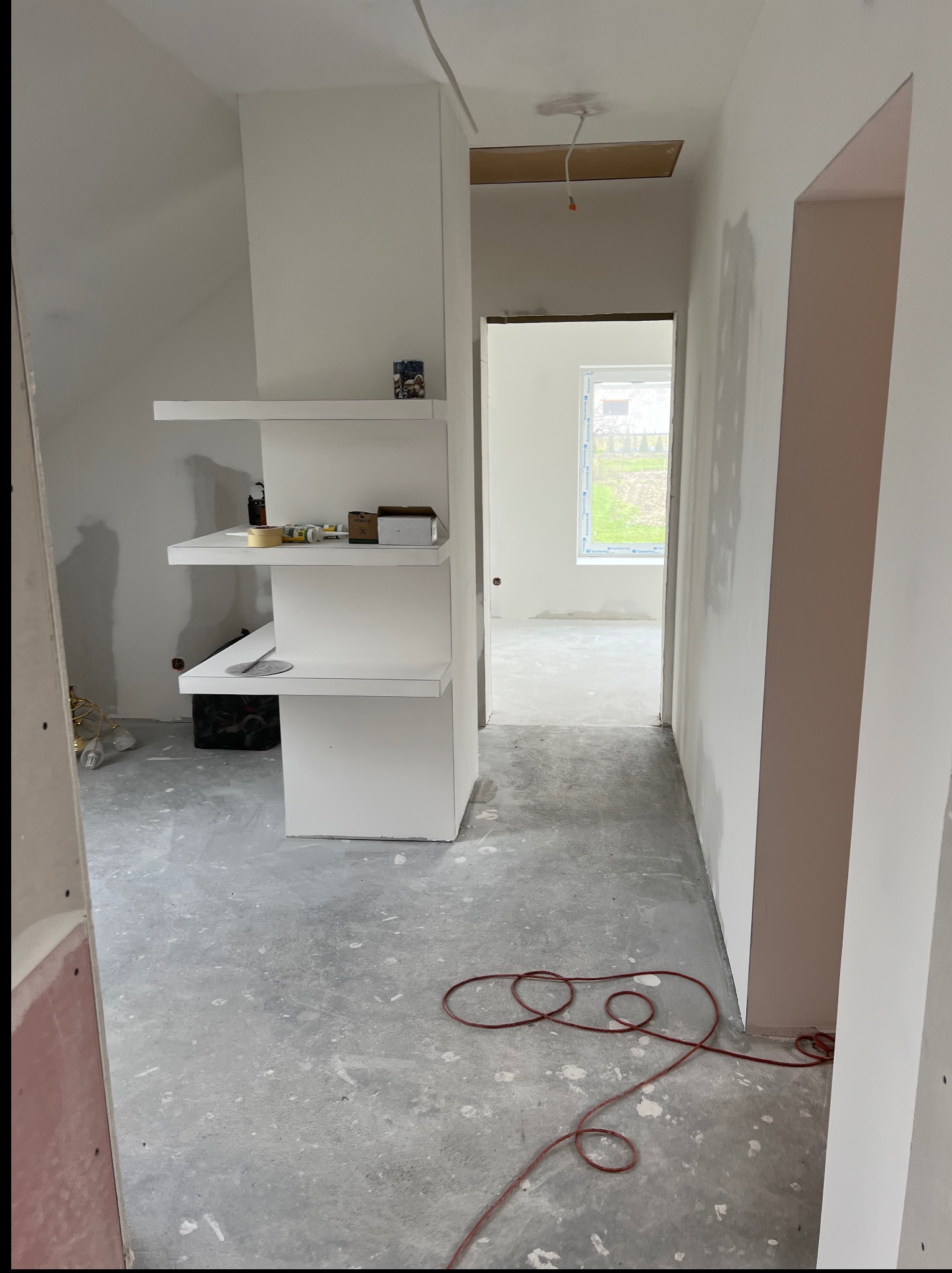 Wnętrze w trakcie wykończenia: surowy beton na podłodze, białe ściany po gładzi, regał z półkami. Widoczny kabel na podłodze i okno w oddali.
