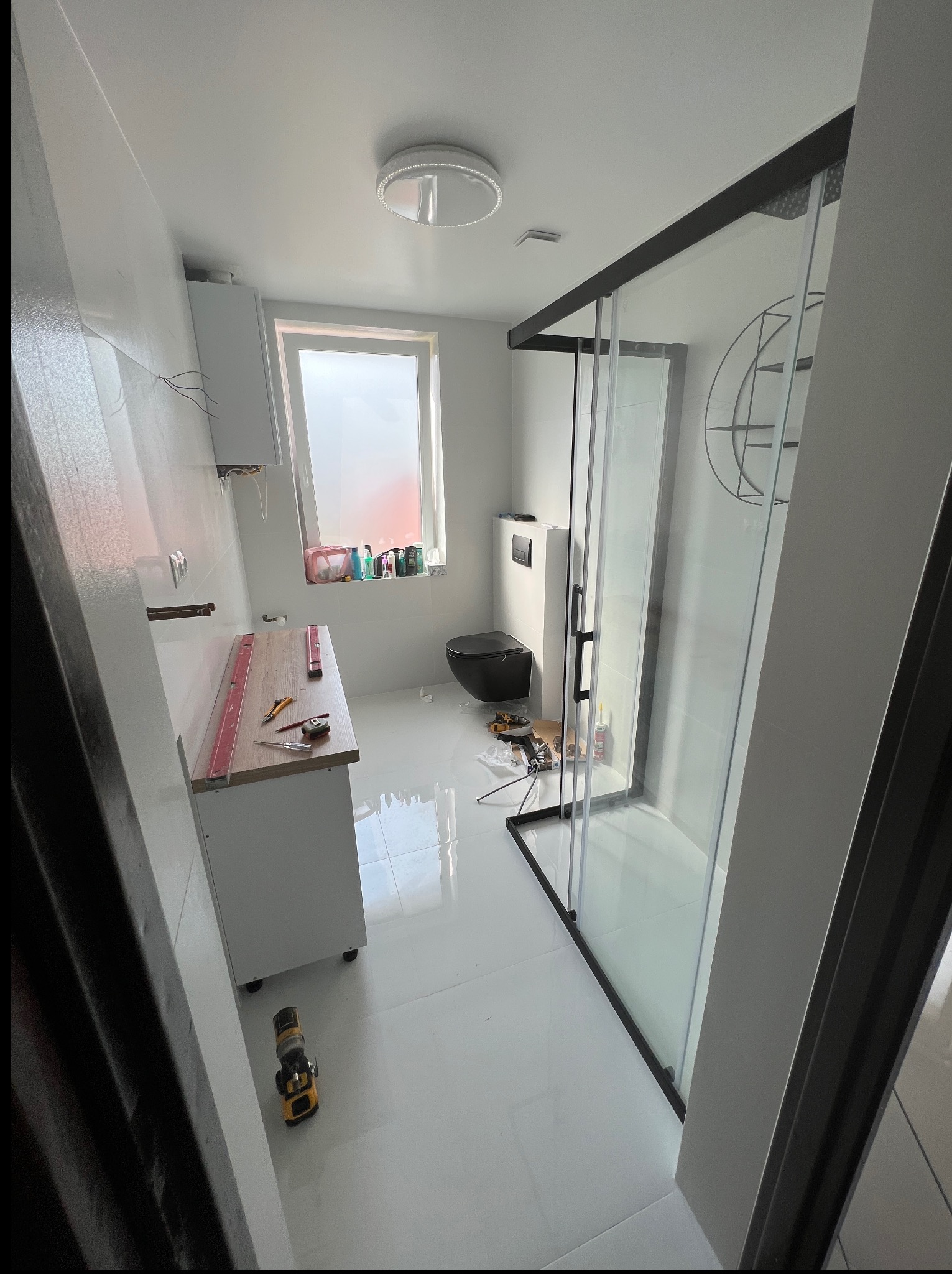 Biała łazienka w trakcie wykańczania: kabina prysznicowa z czarnymi ramami, umywalka z blatem, czarna toaleta. Widoczne narzędzia i poziomice.