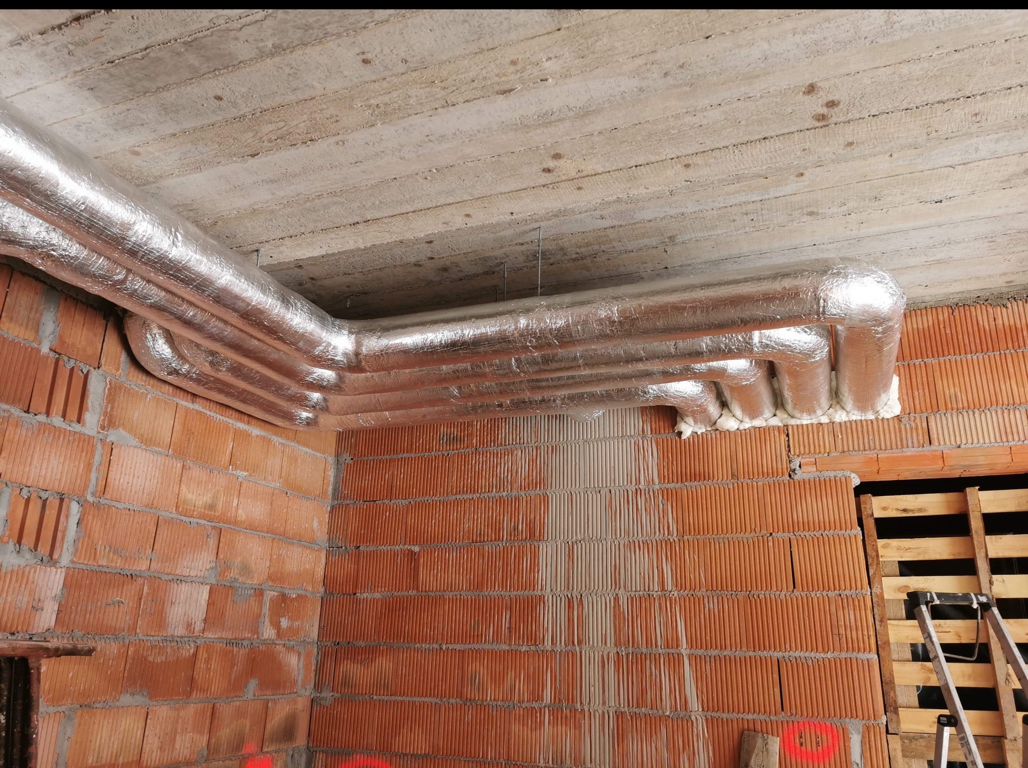 Izolowane rury wentylacyjne pod betonowym stropem w surowym wnętrzu. Widoczne ceglane ściany i drabina. Detal z budowy, instalacja wentylacyjna w trakcie prac.