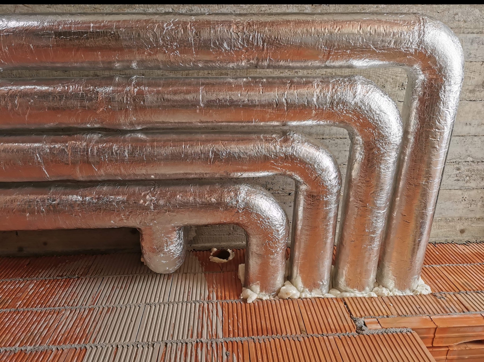 Ocieplone, srebrną folią rury grzewcze, przymocowane pianką montażową do sufitu i podłogi z czerwonej cegły dziurawki. Widoczne załamania rur.