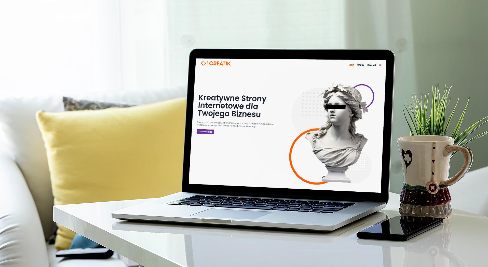 Laptop wyświetlający stronę internetową z motywem klasycznej rzeźby, obok kubek w stylu vintage i smartfon na białym stole.