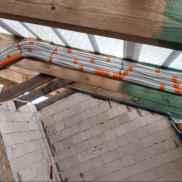 HEGEMON Sp. z o.o. - Kable elektryczne ułożone równolegle na drewnianej konstrukcji dachu, zabezpieczone pomarańczowymi opaskami, widoczne białe ściany z pustaków.