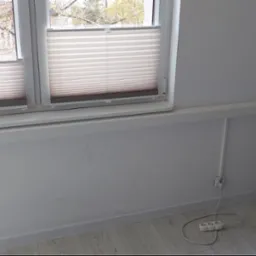 Białe rolety plisowane w oknie, zasłaniające widok drzew bez liści, z maskownicą instalacji pod parapetem i przedłużaczem leżącym na podłodze.