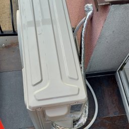 HEGEMON Sp. z o.o. - Zewnętrzna jednostka klimatyzacji zamontowana na balkonie, widoczne przewody zabezpieczone folią aluminiową, fragment balustrady i stopy osoby fotografującej w sportowych butach.