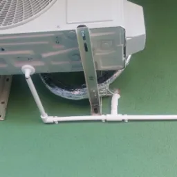 Zewnętrzna jednostka klimatyzatora zamontowana na zielonej ścianie budynku, widoczne białe rury odprowadzające skropliny i srebrna rura chłodnicza.