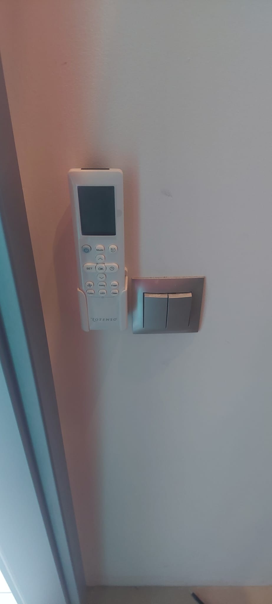 Biały pilot do klimatyzacji marki Rotenso zamocowany na ścianie obok podwójnego włącznika światła z szarą ramką.