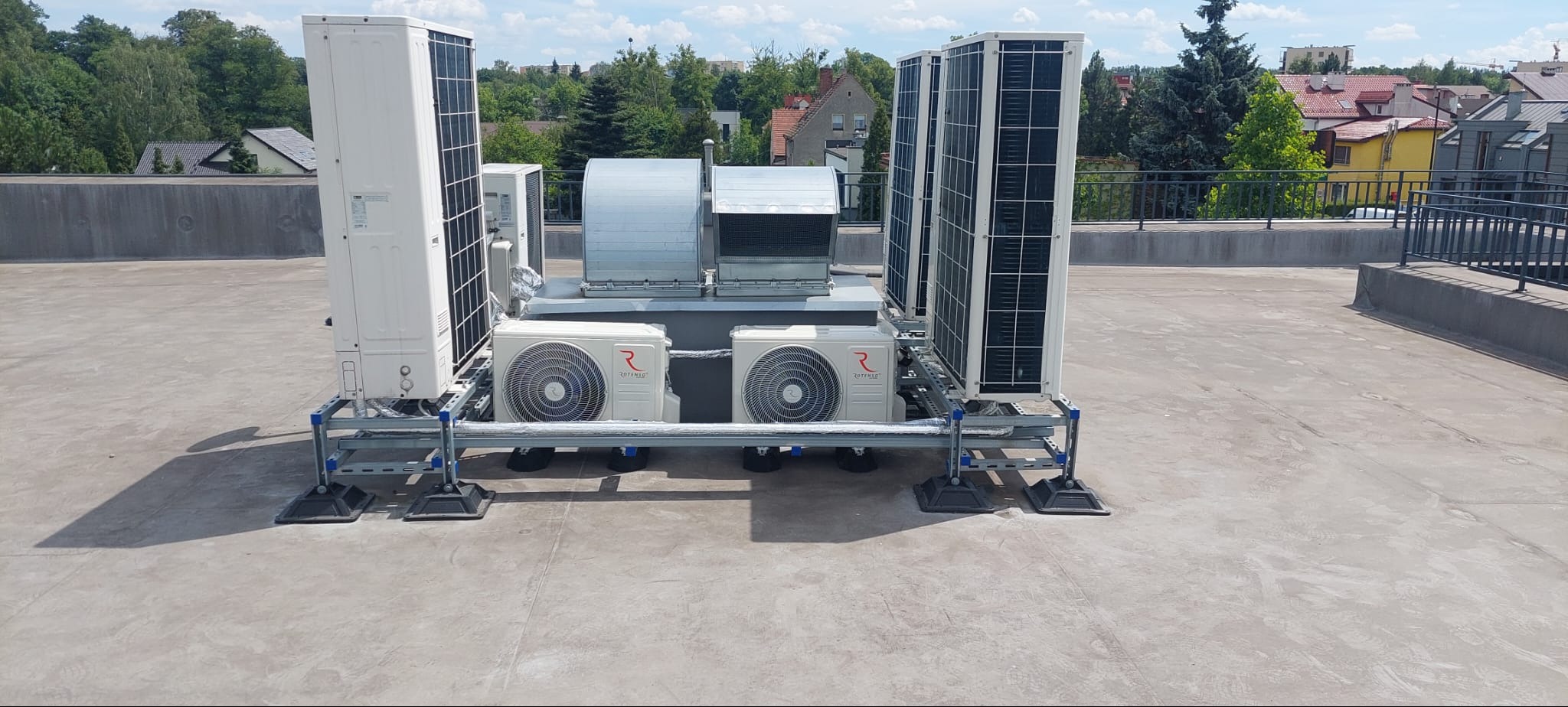 Zestaw urządzeń klimatyzacyjnych i wentylacyjnych na dachu budynku, widoczne jednostki zewnętrzne klimatyzatorów, kanały wentylacyjne oraz metalowa konstrukcja wsporcza.