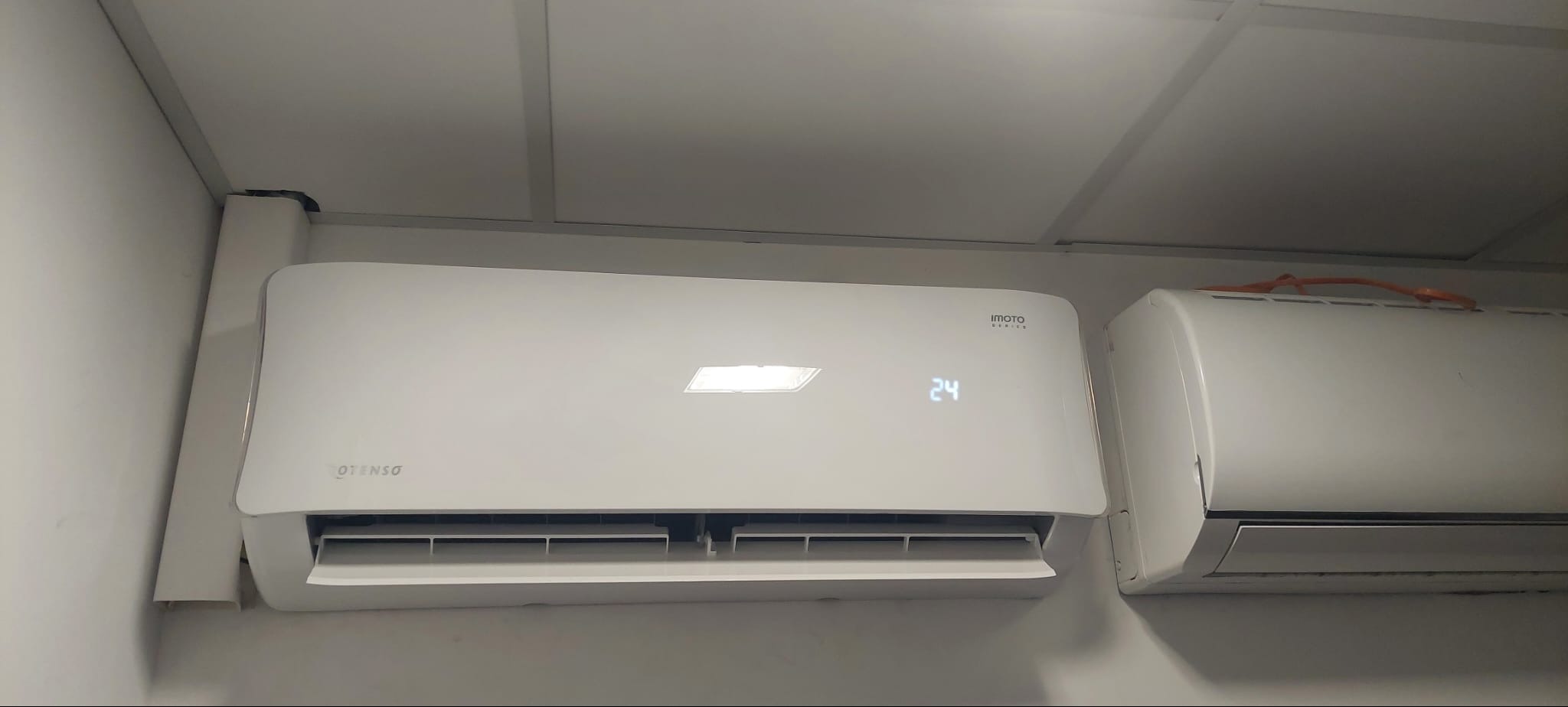 Biały klimatyzator ścienny marki Imoto z wyświetlaczem wskazującym 24 stopnie, zamontowany obok drugiego klimatyzatora i maskownicy przewodów.