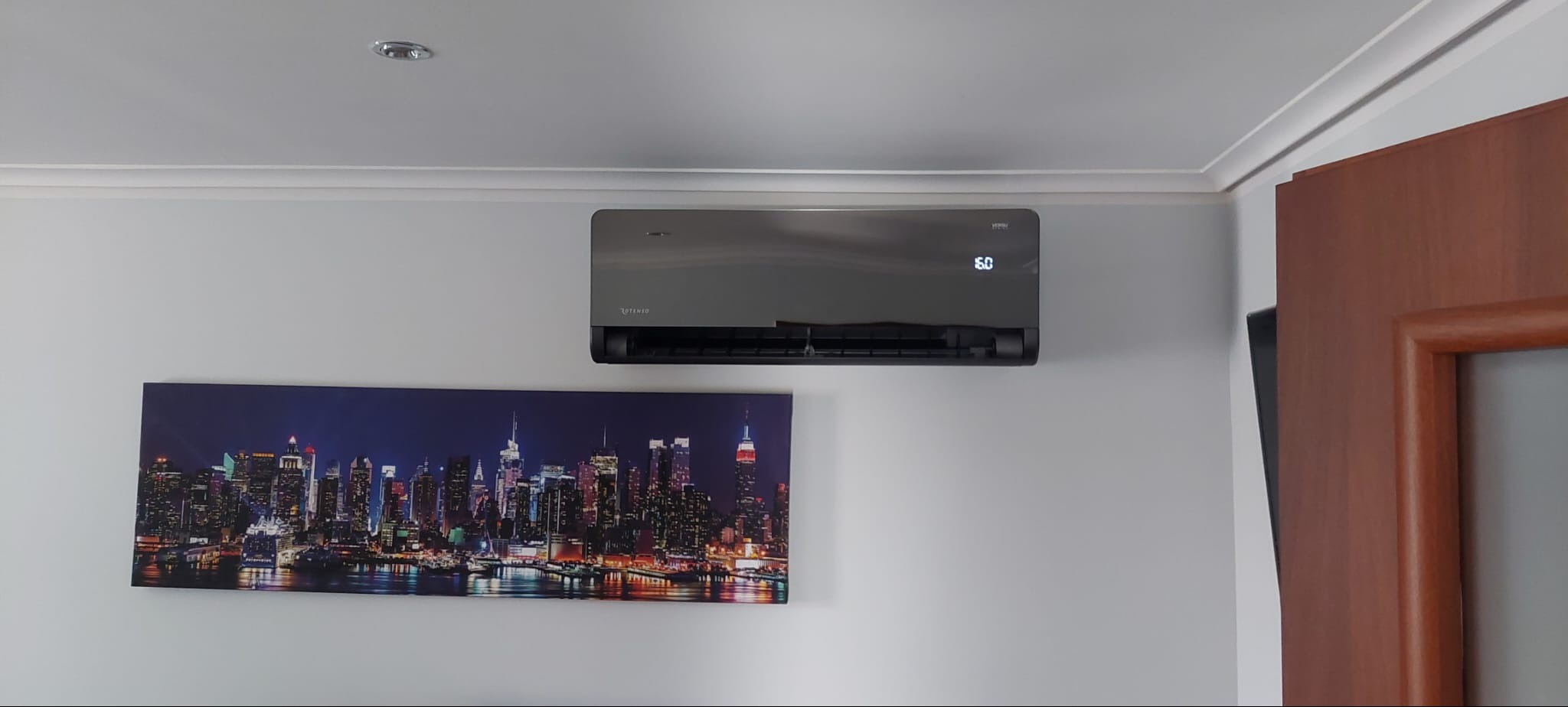 Szary klimatyzator ścienny z wyświetlaczem temperatury 16 stopni Celsjusza, zamontowany nad podświetlanym obrazem panoramy miasta nocą.