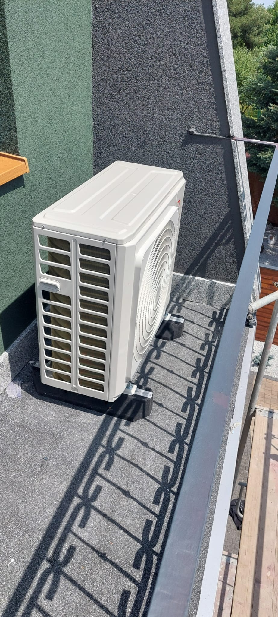 Zewnętrzna jednostka klimatyzacji zamontowana na dachu, widoczne gumowe podkładki antywibracyjne i cień barierki.