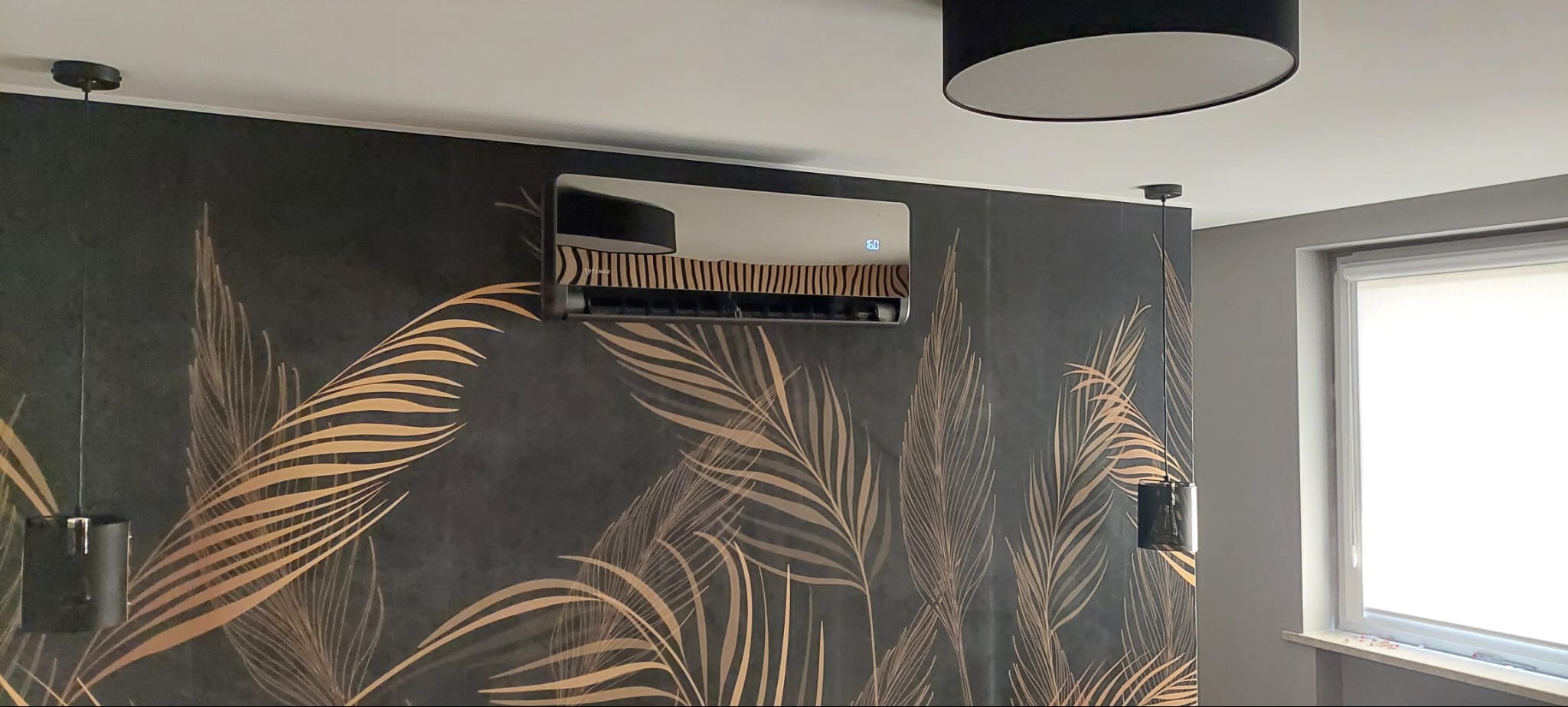 Czarny klimatyzator ścienny zamontowany na ścianie z motywem liści palmowych, widoczny wyświetlacz temperatury, oświetlenie sufitowe w tle.