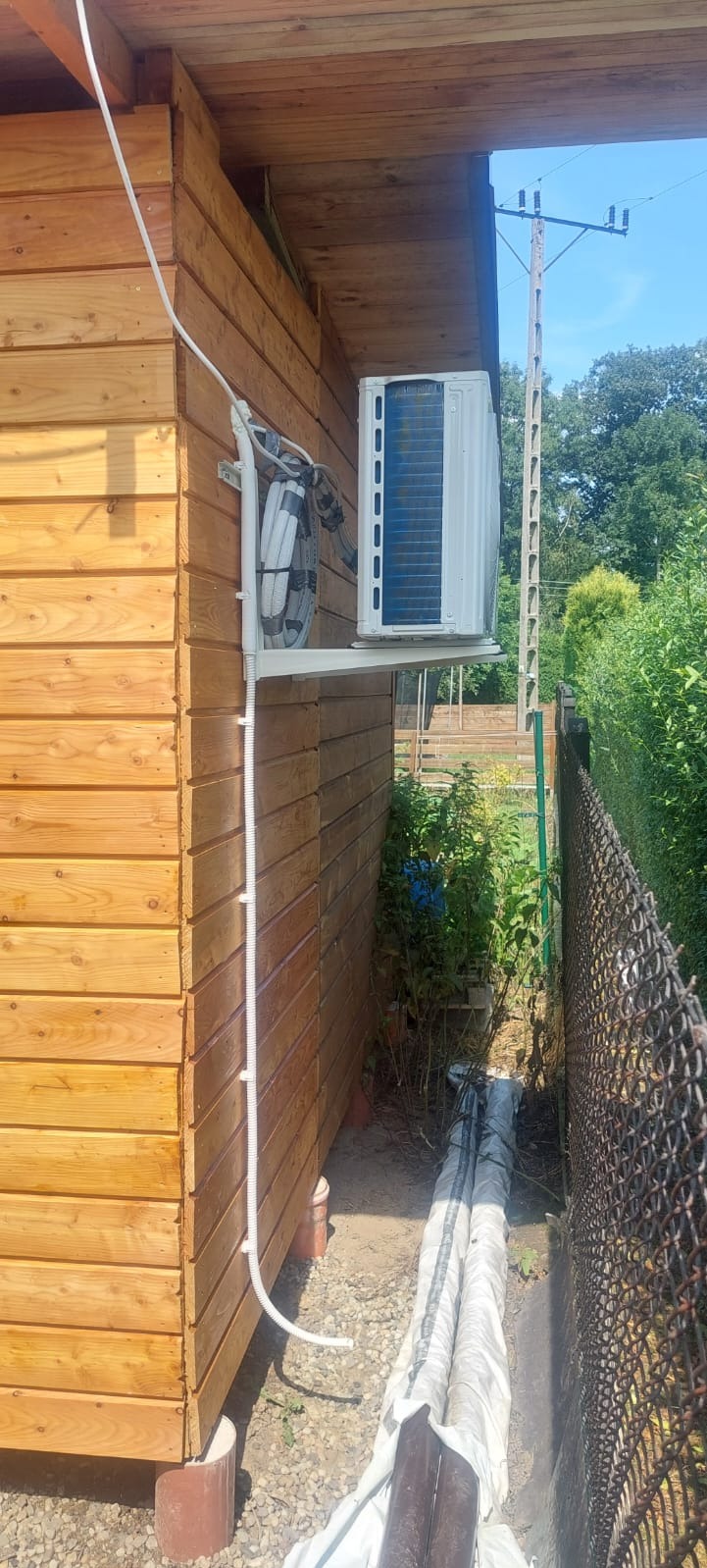Biała jednostka zewnętrzna klimatyzacji zamontowana na drewnianej ścianie budynku, z widocznymi przewodami i wspornikiem montażowym, obok zarośnięty teren i siatka ogrodzeniowa.