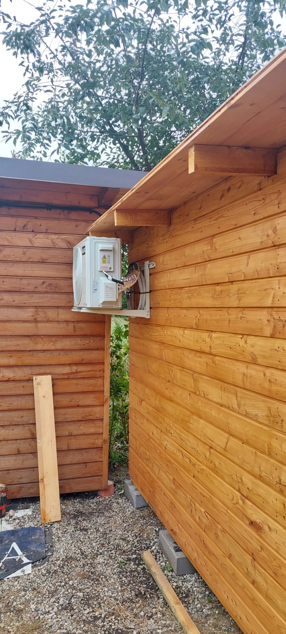 Zewnętrzna jednostka klimatyzacji zamontowana na drewnianej ścianie budynku gospodarczego, widoczne przewody w osłonie i elementy montażowe, tło stanowią drzewa i żwirowe podłoże.