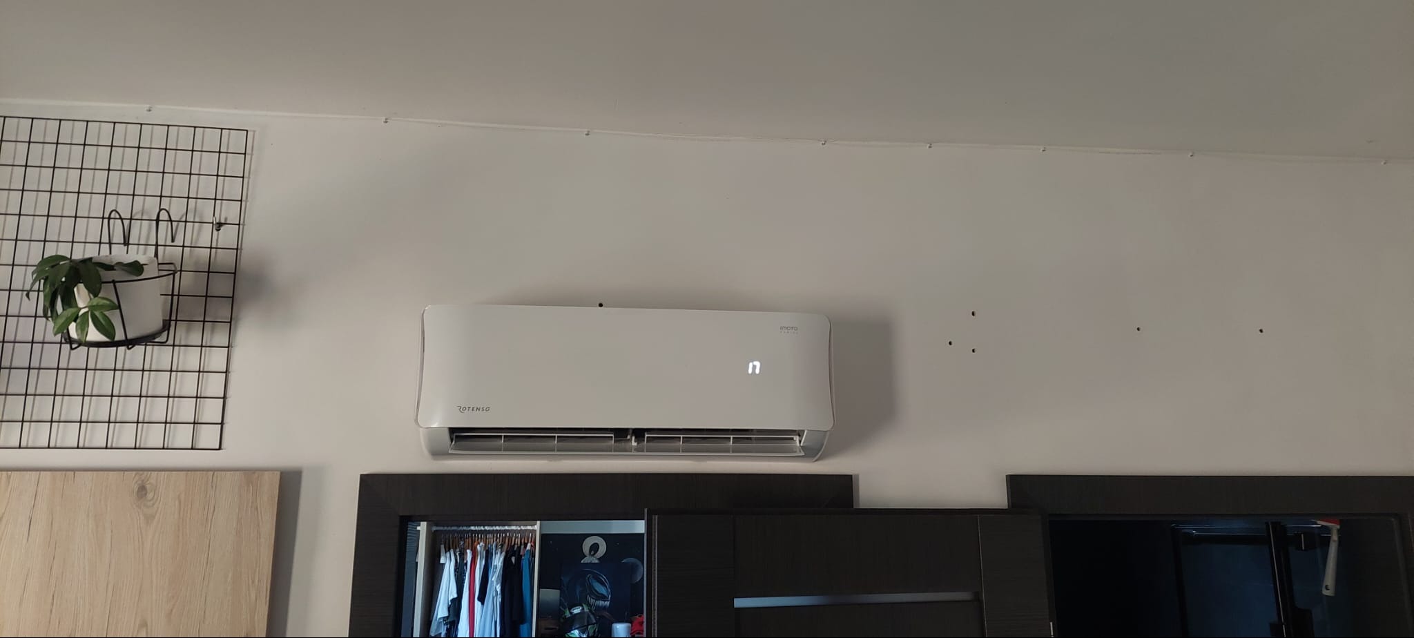 Biały klimatyzator marki Rotenso zamontowany na ścianie nad szafkami, z widocznym wyświetlaczem temperatury i czarną kratką z doniczką po lewej stronie.