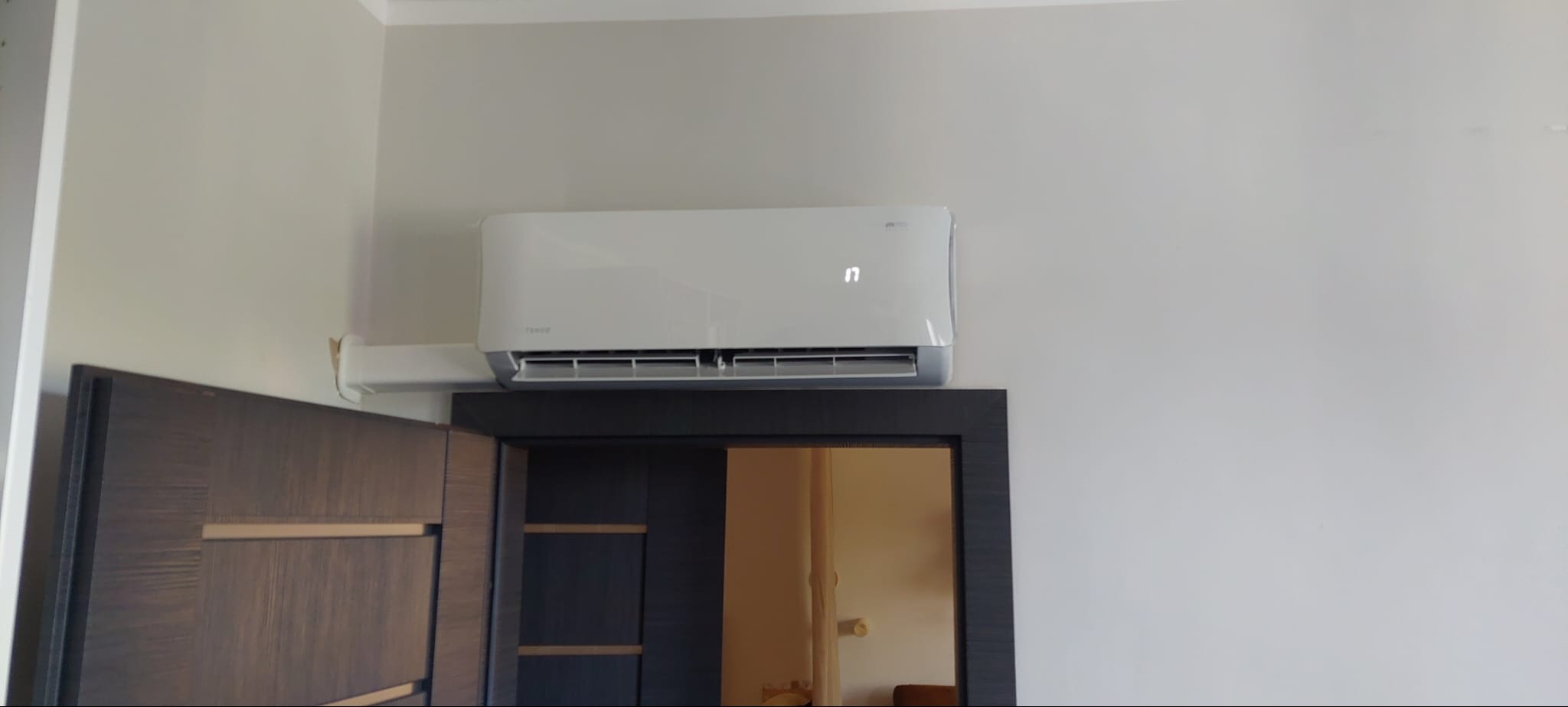 Biały klimatyzator na ścianie nad ciemną, drewnianą ramą drzwi, widoczny wyświetlacz temperatury.