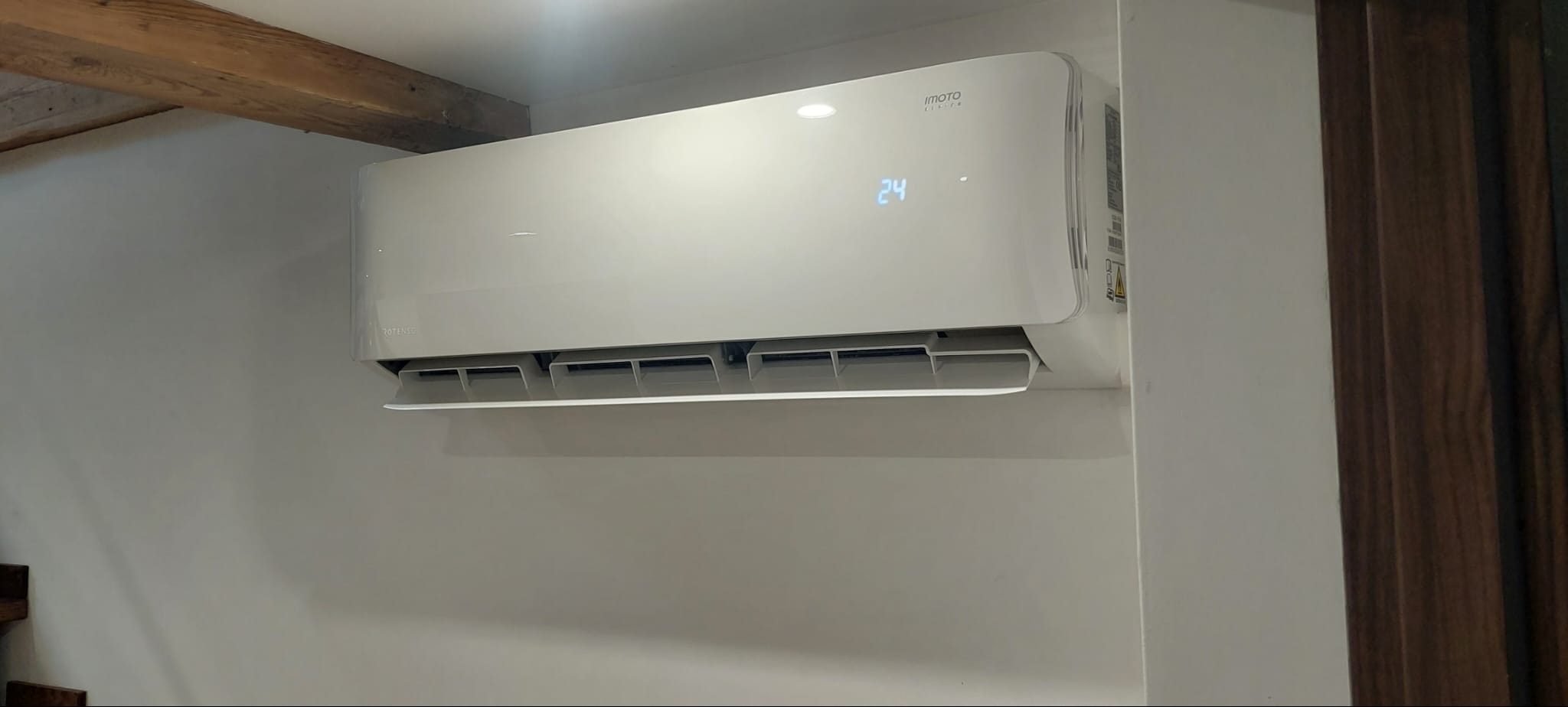 Biały klimatyzator marki IMOTO zamontowany na ścianie, wyświetlający temperaturę 24 stopnie, widoczny fragment drewnianego stropu i schodów.