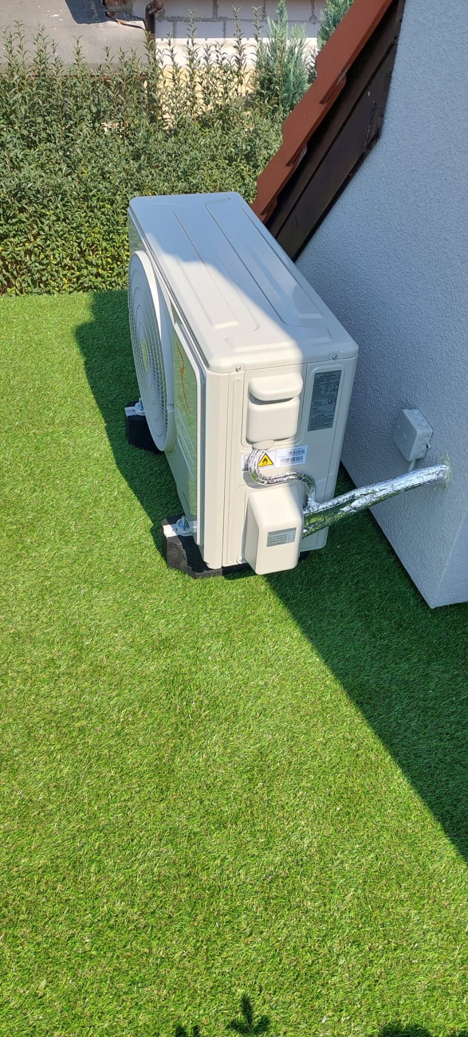 Zewnętrzna jednostka klimatyzacji zamontowana na trawie syntetycznej przy ścianie budynku, z widocznymi przewodami w izolacji aluminiowej.