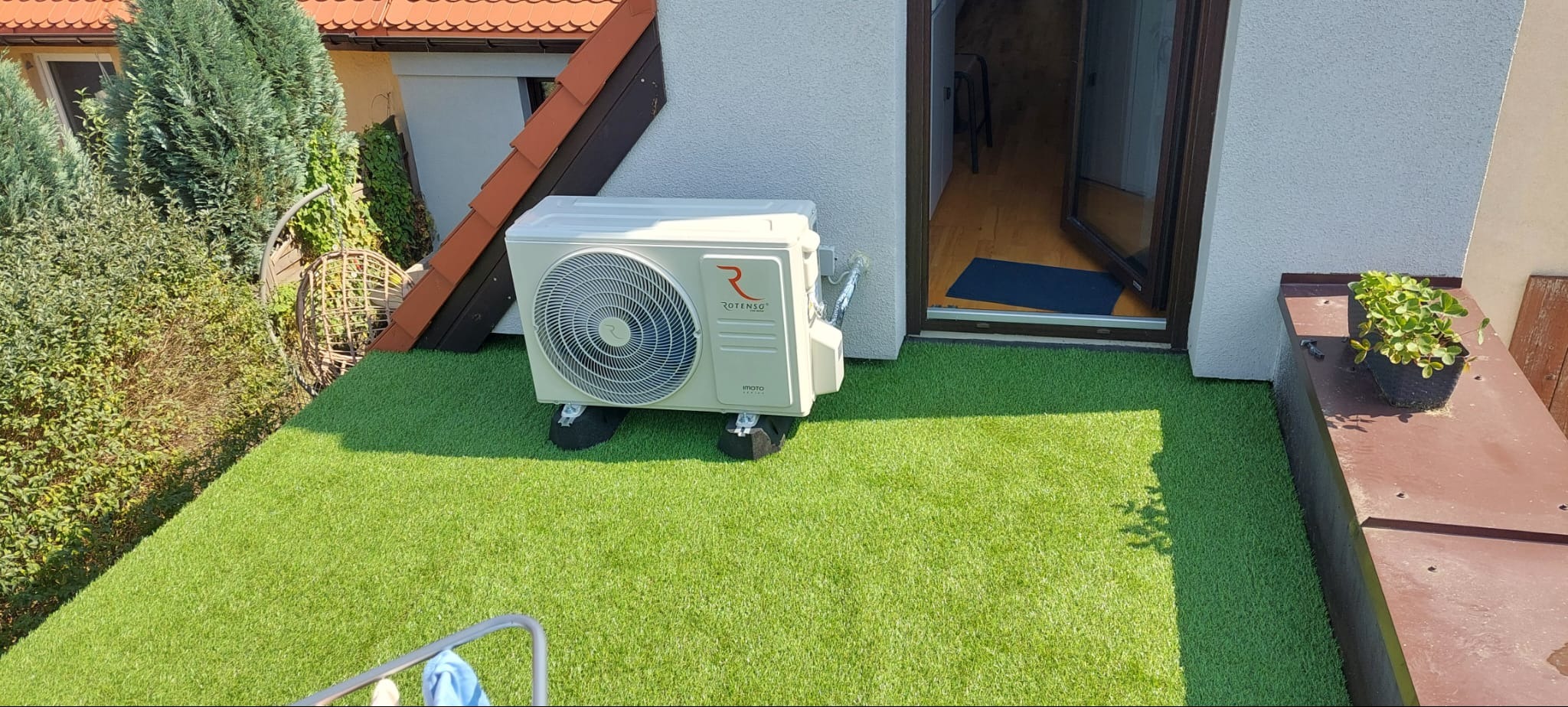 Zewnętrzna jednostka klimatyzacji Rotenso na dachu pokrytym sztuczną trawą, widok na otwarte drzwi balkonowe i rośliny w doniczce.