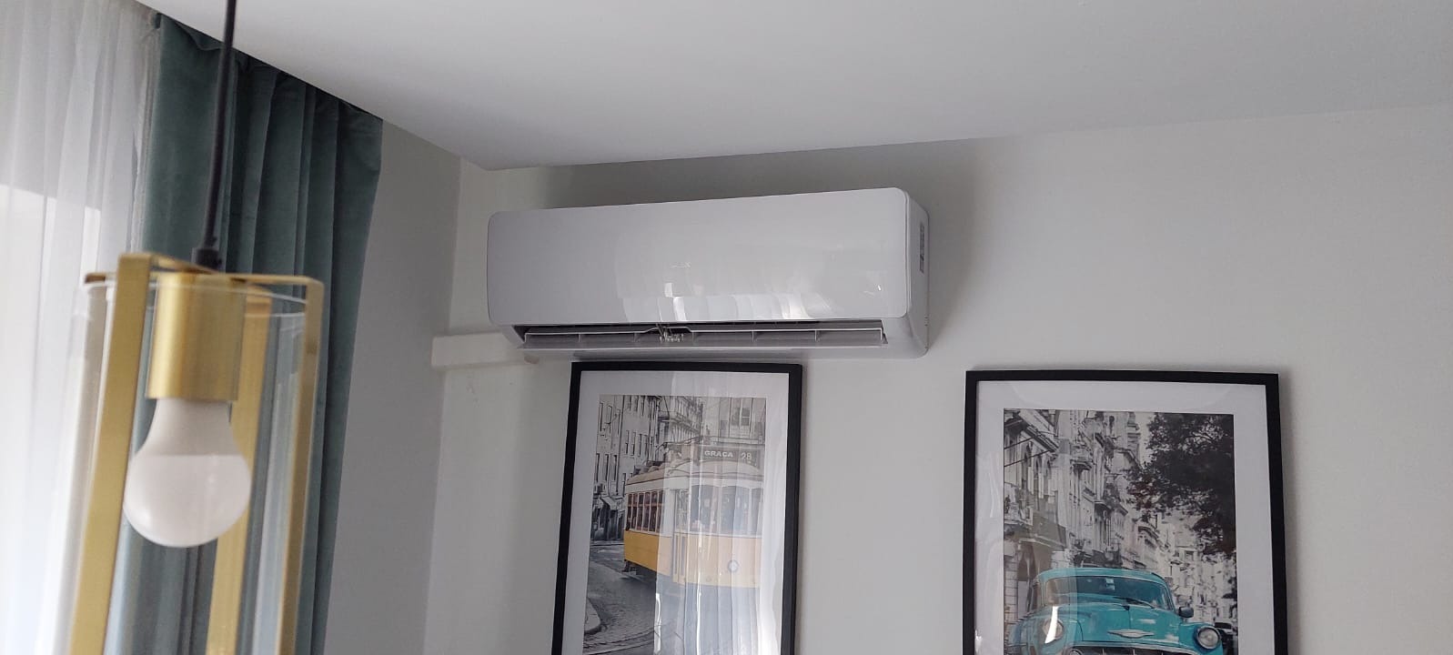 Biały klimatyzator ścienny zamontowany nad dwoma oprawionymi w ramki grafikami przedstawiającymi miejskie sceny, oświetlony wiszącą lampą z elementami w kolorze złota i zasłoną w tle.