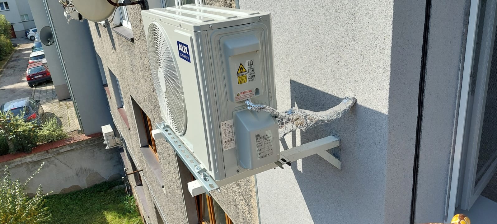 Zewnętrzna jednostka klimatyzacji marki AUX zamontowana na elewacji budynku, widoczne rury w izolacji aluminiowej oraz uchwyty montażowe.