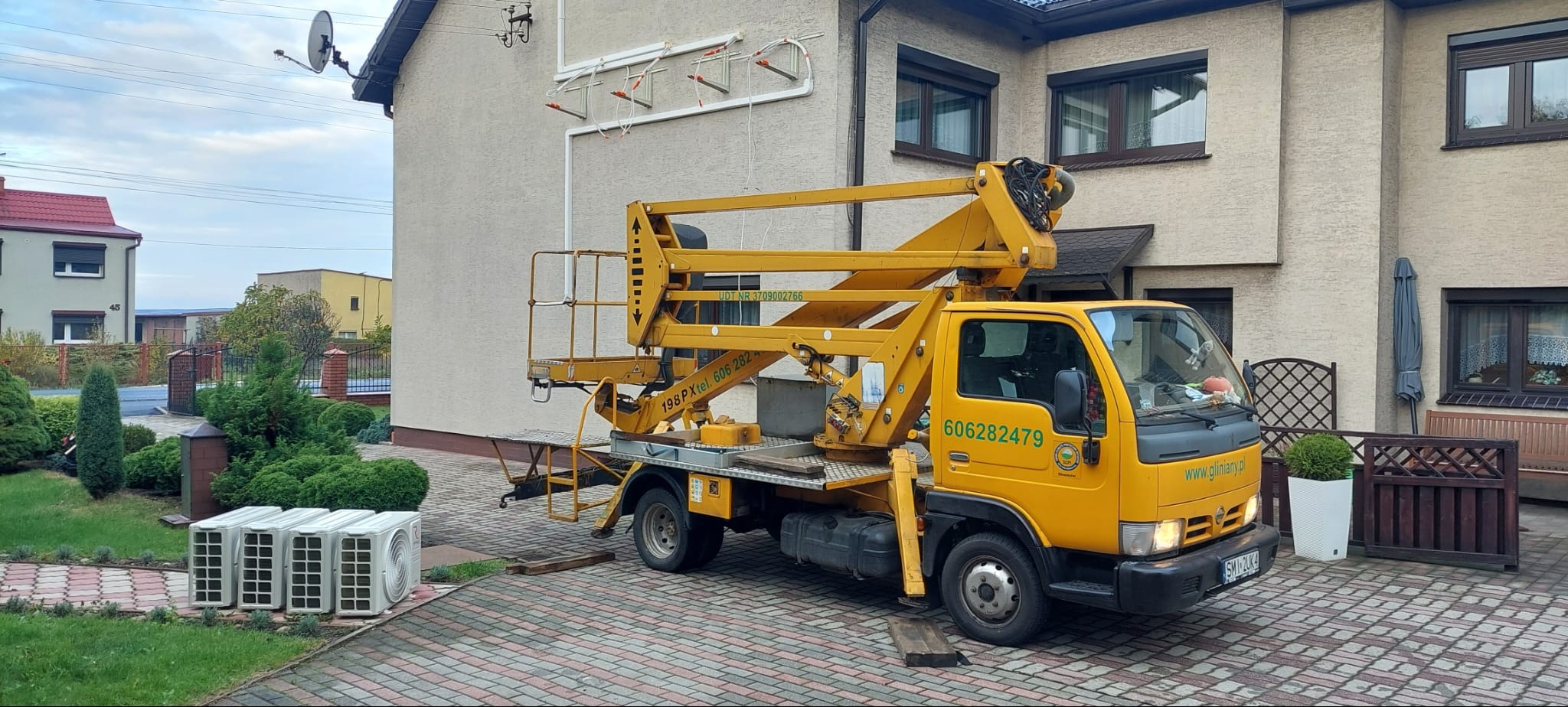 Żółty podnośnik koszowy zaparkowany na posesji przed domem, obok leżą cztery jednostki zewnętrzne klimatyzacji, na ścianie domu widoczne rury instalacji.