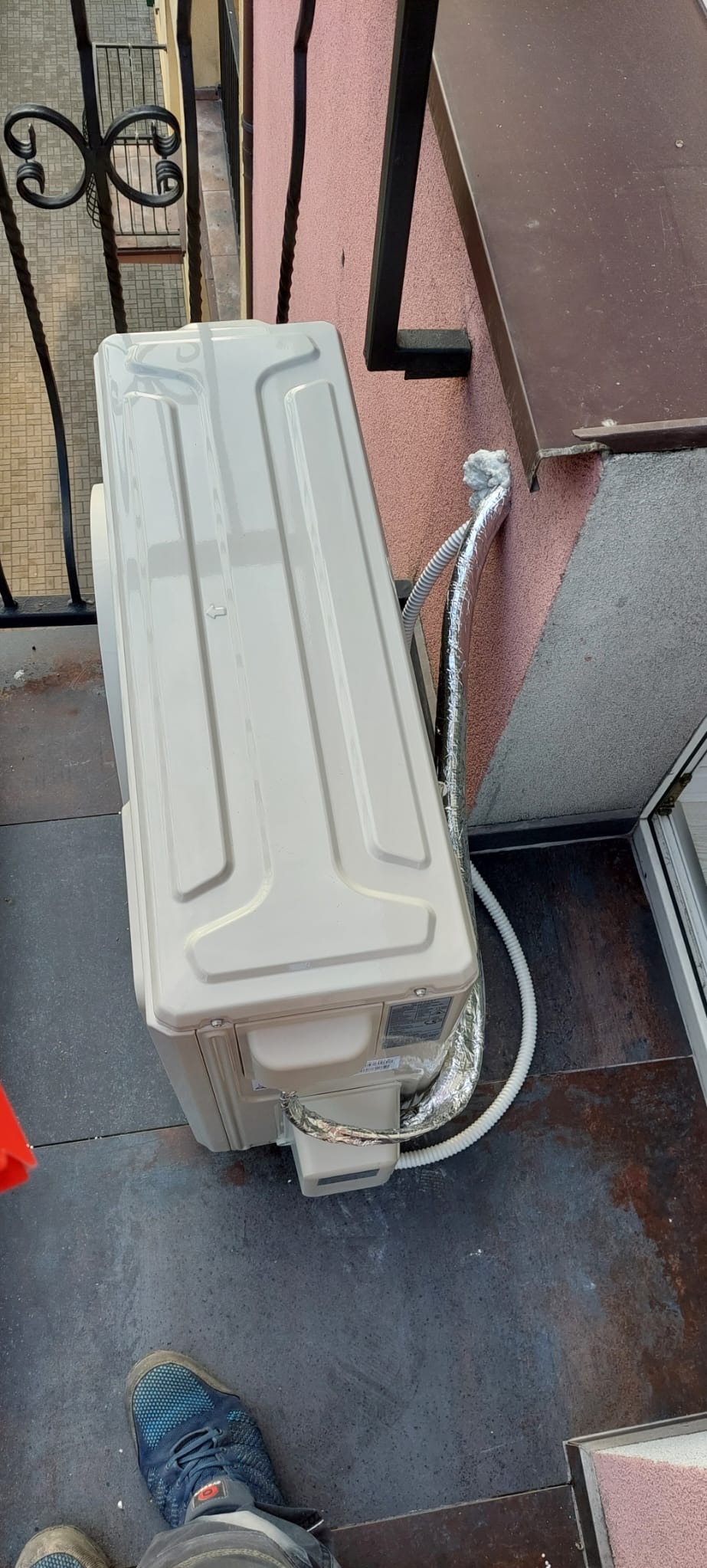 Zewnętrzna jednostka klimatyzacji zamontowana na balkonie, widoczne przewody zabezpieczone folią aluminiową, fragment balustrady i stopy osoby fotografującej w sportowych butach.