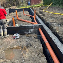 ArtM-Bud - Budowa fundamentów z bloczków betonowych, widoczne pomarańczowe rury kanalizacyjne w gruncie i pracownik w czerwonej kurtce.