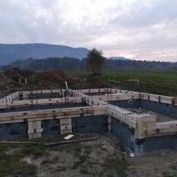 ArtM-Bud - Wznoszenie fundamentów domu z bloczków betonowych, widoczne szalunki drewniane, zbrojenie pionowe, w tle hałda ziemi i zalesione wzgórza pod pochmurnym niebem.