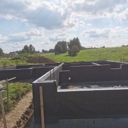 ArtM-Bud - Zabezpieczone fundamenty domu jednorodzinnego z widoczną izolacją pionową, na tle zieleni i błękitnego nieba z chmurami.