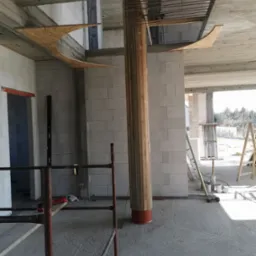 Wnętrze w stanie surowym: murowane ściany z betonu komórkowego, drewniana kolumna w szalunku, fragment deskowania na suficie, rusztowanie i drabiny w tle.