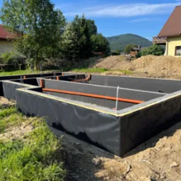 Zabezpieczone fundamenty z izolacją pionową i poziomą, wystające rury kanalizacyjne. W tle domy i drzewa w słoneczny dzień.