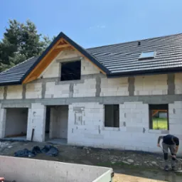Surowy stan domu z białych bloczków, czarny dach z lukarną, pracownik na zewnątrz. Widok na góry w tle. Dom w trakcie budowy.