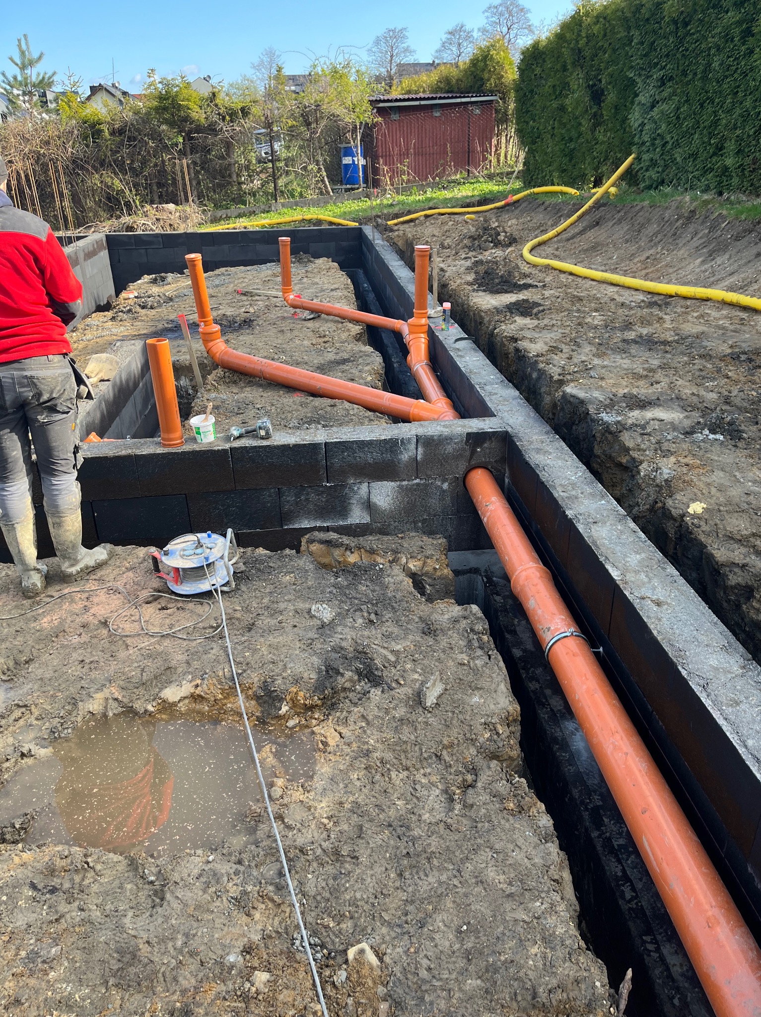 Budowa fundamentów z bloczków betonowych, widoczne pomarańczowe rury kanalizacyjne w gruncie i pracownik w czerwonej kurtce.