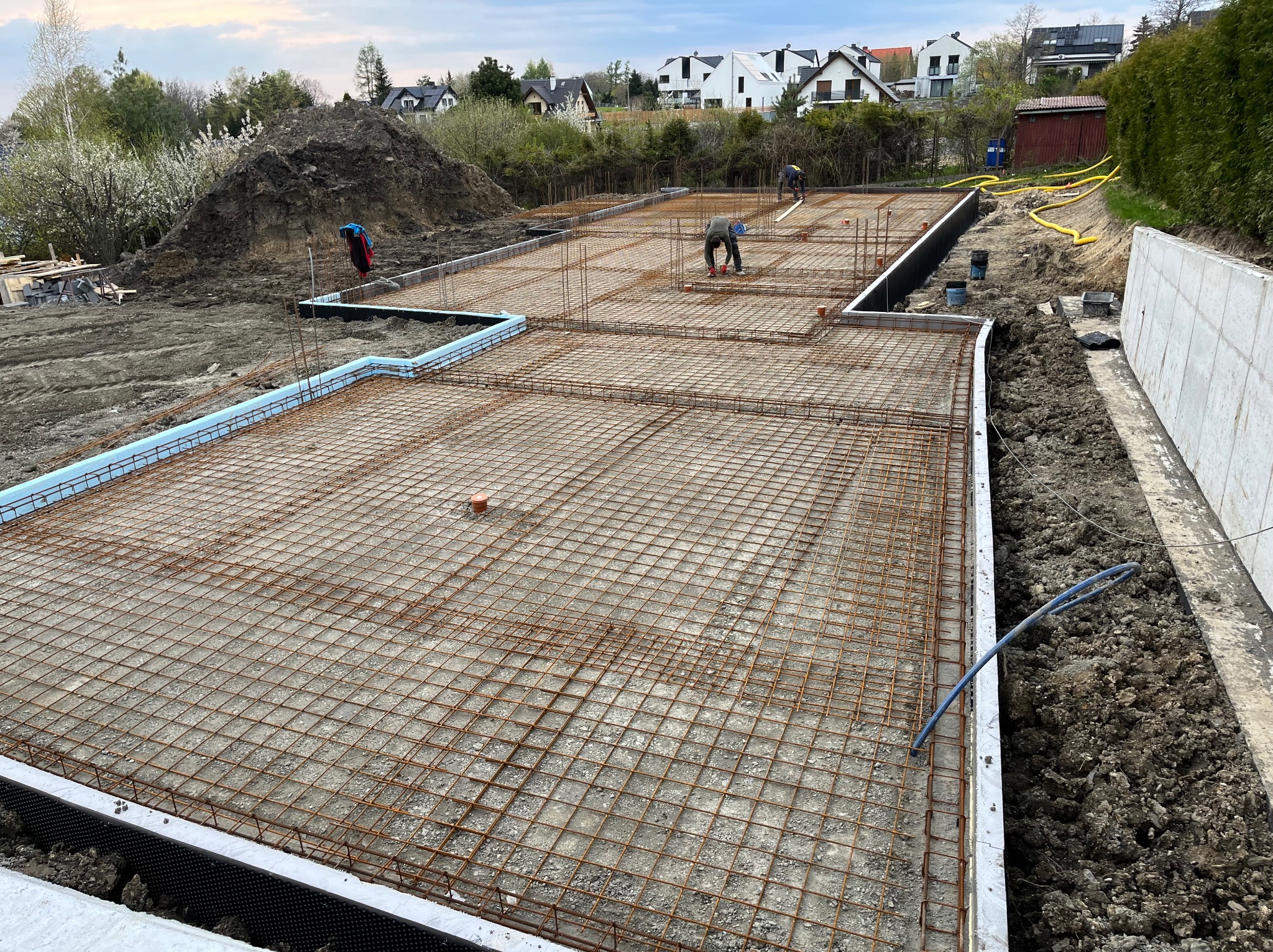 Zbrojenie płyty fundamentowej z widoczną izolacją pionową i poziomą, pracownicy montują zbrojenie na budowie domu jednorodzinnego.