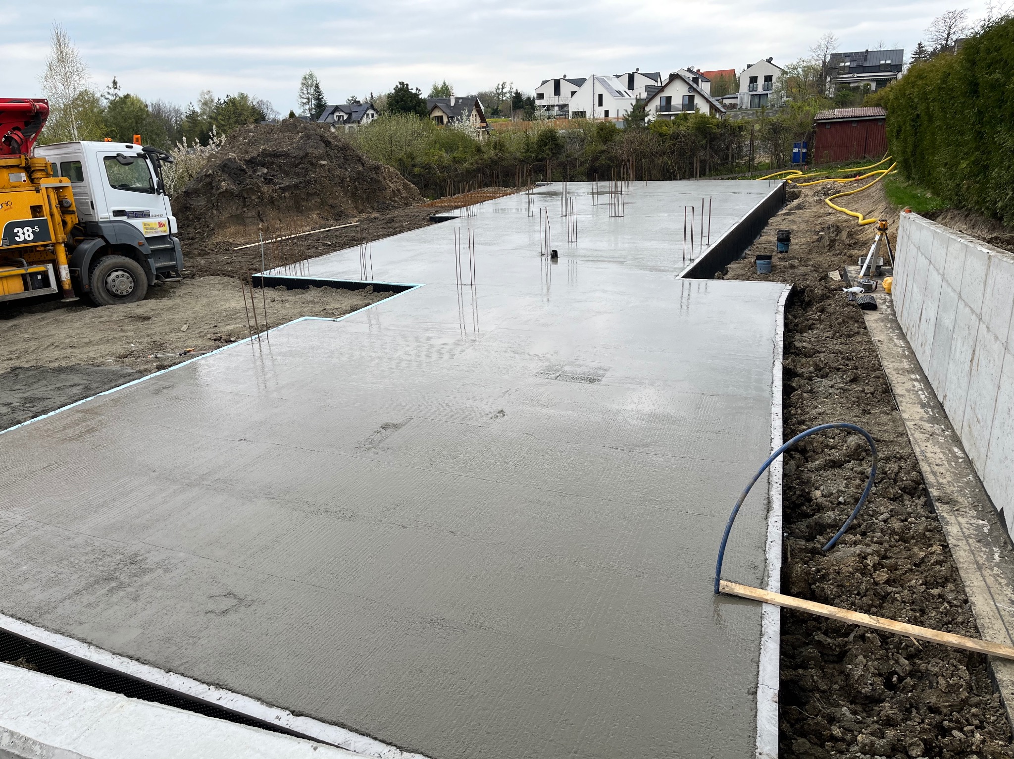 Wylany betonowy fundament domu jednorodzinnego z wystającymi prętami zbrojeniowymi, widoczna pompa do betonu i pryzma ziemi w tle zabudowy mieszkalnej.