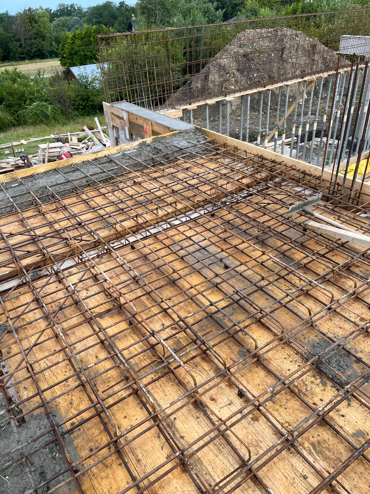 Zbrojenie stropu ułożone na deskowaniu, częściowo zalane betonem, widok na zewnątrz na teren budowy z ziemią i konstrukcjami wsporczymi.