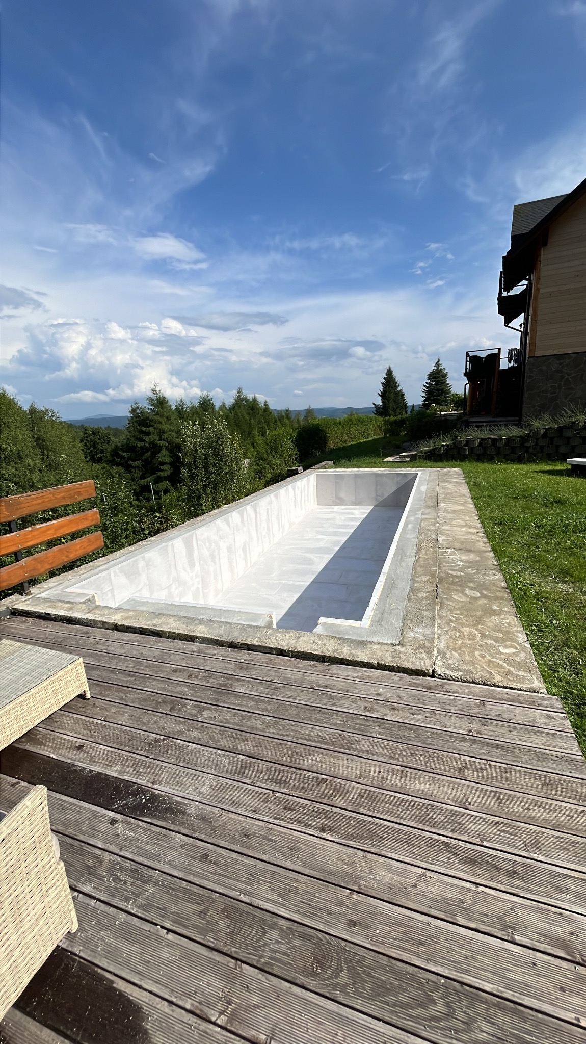Niedokończony betonowy basen w ziemi na drewnianym tarasie, z widokiem na zielone wzgórza i błękitne niebo z chmurami w tle.