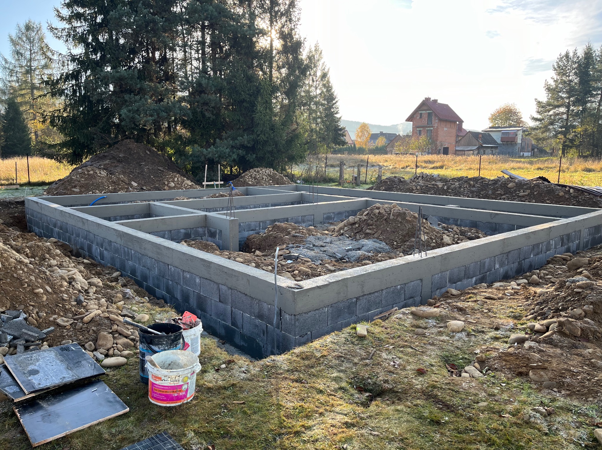 Wylane fundamenty z bloczków betonowych i betonu, widoczne zbrojenia, teren budowy z hałdami ziemi i narzędziami, w tle dom w stanie surowym otwartym i drzewa.