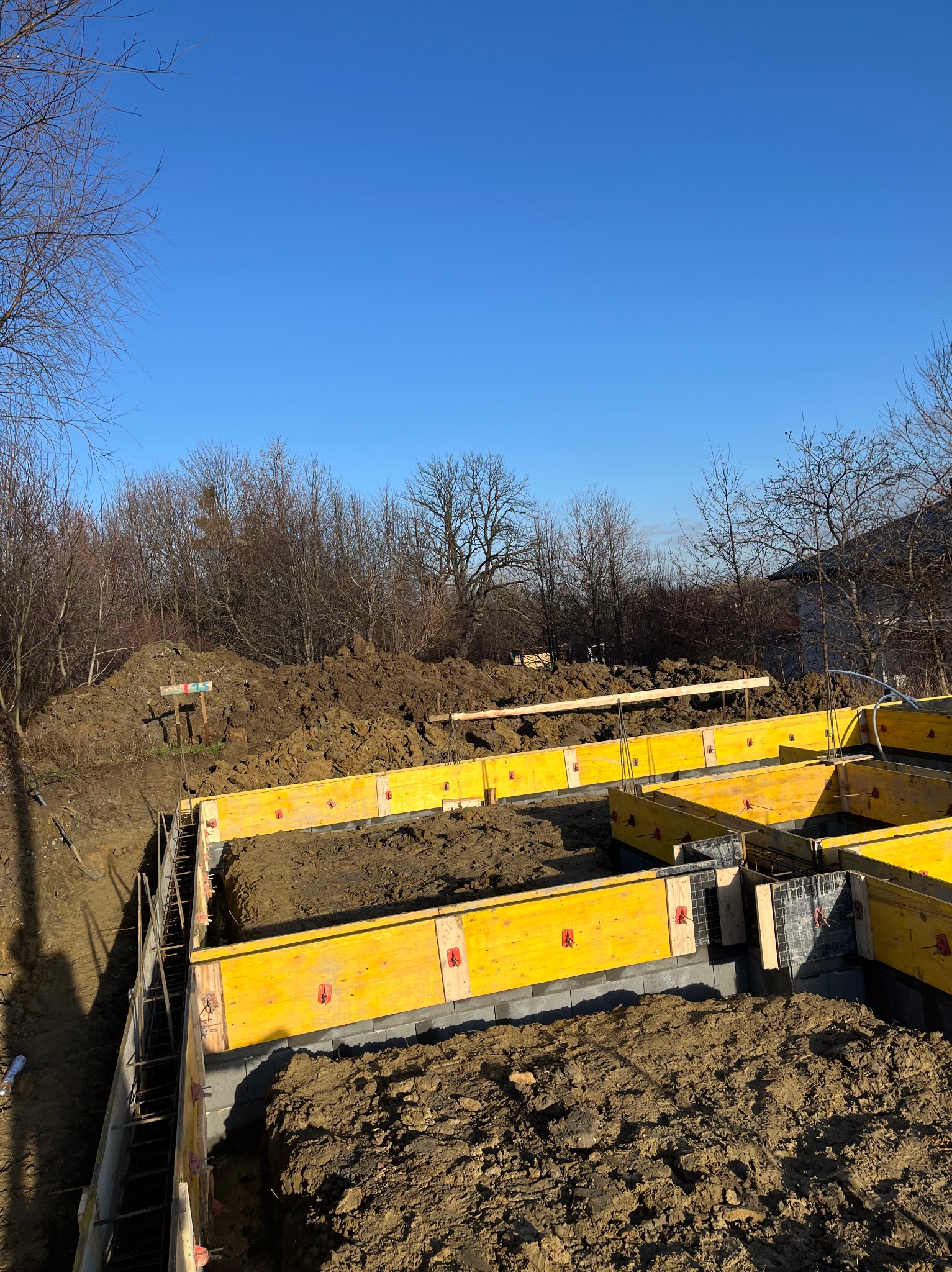 Żółte szalunki ustawione w wykopie, przygotowane do zalewania betonem, widoczne zbrojenia i fragment domu w tle.