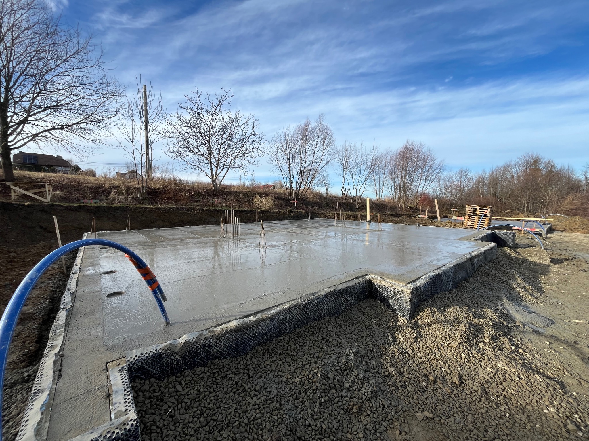 Świeżo wylana płyta fundamentowa z widocznymi prętami zbrojeniowymi, zabezpieczona folią kubełkową, z niebieskim przewodem wystającym z betonu, na tle błękitnego nieba z chmurami i drzew bez liści.