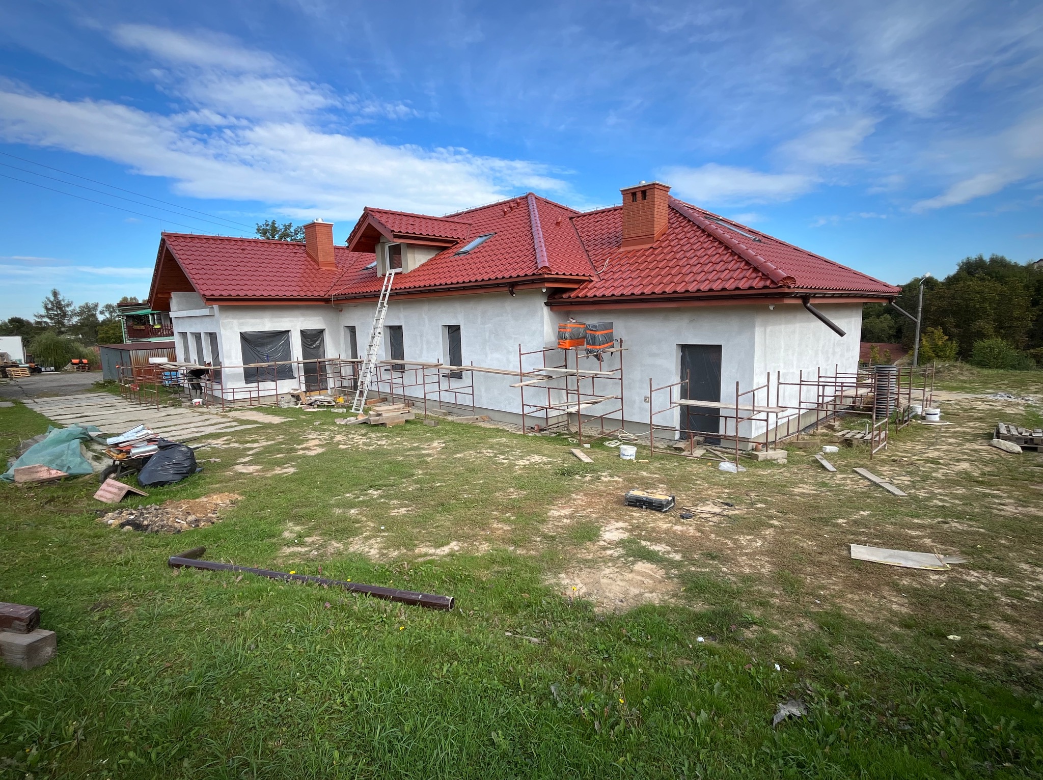 Budowa domu murowanego z czerwoną dachówką, widoczne rusztowania, częściowo otynkowana elewacja i przygotowany teren wokół budynku.